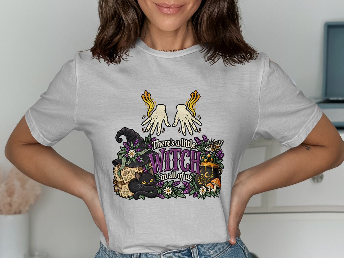 White t-shirt with a graphic featuring a black cat, witch hat, cauldron, and text "There's a little WITCH in all of us."